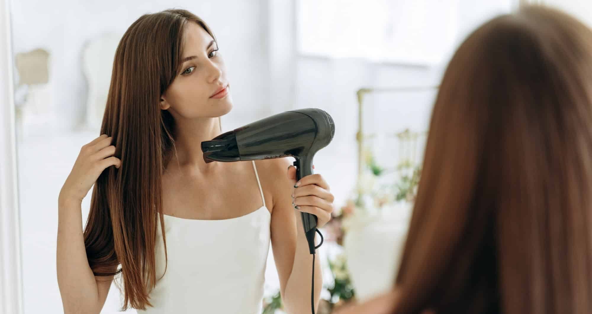 Beautiful girl using a hair dryer and smiling while looking at the mirror.