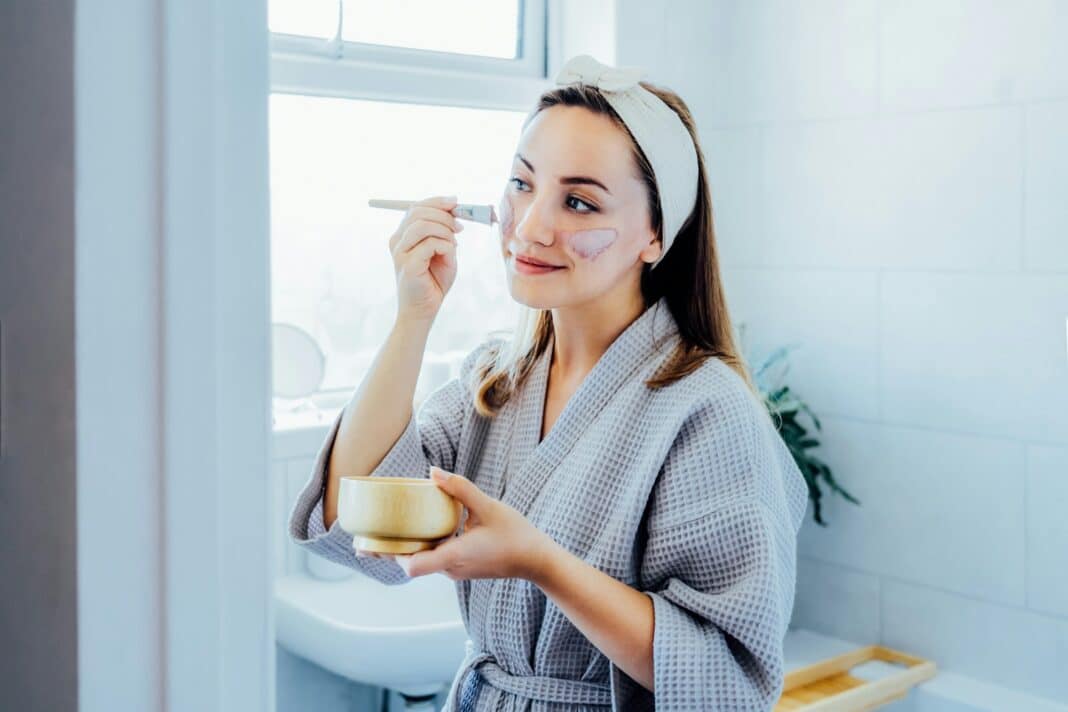 Young woman in bathrobe looking in the mirror and applying facial natural cosmetic clay mask on face